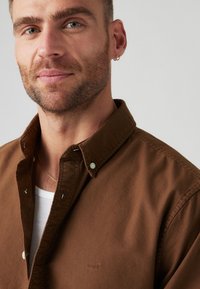 Brown button-up shirt with a collar, featuring white buttons and subtle Levi's logo. Textured fabric; model has short hair and a light beard.