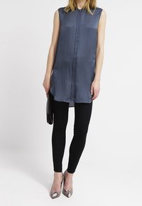 Ärmellose, knöpfbare Tunika aus schiefergrauem Stoff, kombiniert mit schwarzen Leggings und metallischen spitzen Absatzschuhen. Schwarze Clutch in der Hand.