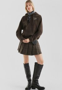 Młoda kobieta ubrana w brązowy hoodie, w kratkowaną spódnicę plisowaną, czarne botki i czarny wzorzysty szalik przewiązany wokół szyi.
