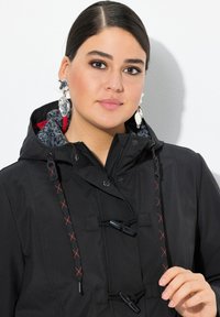 Veste à capuche noire avec doublure grise texturée, accents à carreaux rouges et fermetures à bascule. Caractéristiques de cordons de serrage réglables avec couture rouge.