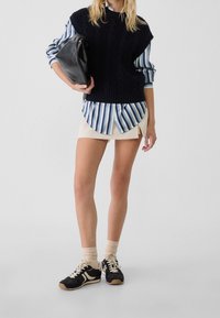 Kvinna klädd i randig blå skjorta under svart stickad väst, beige minikjol, svart och vit sneakers, håller en stor svart handväska.