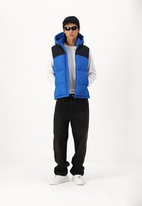 Gilet matelassé bleu et noir avec capuche, porté sur une chemise blanche à manches longues ; associé à un pantalon noir et des baskets blanches.