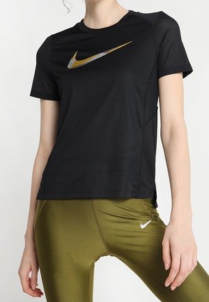Mujer vistiendo una camiseta negra de manga corta de Nike con el logo Swoosh dorado y leggings verde oliva con el logo blanco de Nike, de pie sobre un fondo liso.