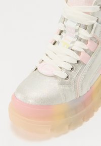 Zapatilla plateada gruesa con acentos rosa pastel, que presenta una tela reflectante, cordones redondos y una suela translúcida con degradado.