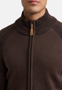 Brun strikket trøje med høj krave, med lynlås og strukturerede sømdetaljer langs skuldrene.