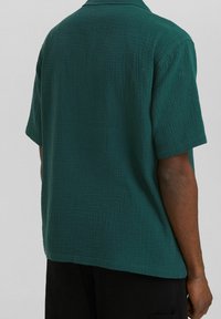 Camicetta a maniche corte in verde scuro, realizzata in un tessuto testurizzato con un sottile motivo a griglia; presenta un colletto classico e una vestibilità rilassata.