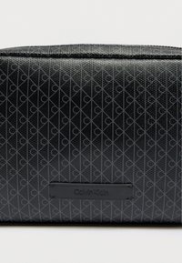 Μαύρο καλλυντικό τσαντάκι με ανάγλυφο σχέδιο από συνδεδεμένα γράμματα και ορθογώνια ετικέτα με το λογότυπο της μάρκας. Κατασκευασμένο από υλικό συνθετικής υφής.