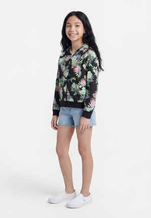 Smiling girl standing, wearing a black floral zip-up hoodie, light blue denim shorts, and white slip-on shoes on a white background.