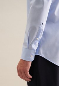 Camicia azzurro chiaro con una texture liscia. Presenta bottoni bianchi e un piccolo design ricamato sul polsino. Vestibilità sartoriale con orlo curvato.