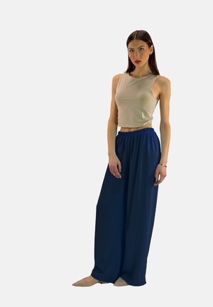 Kobieta w beżowym topie bez rękawów i szerokich granatowych spodniach stojąca na jednolitym tle.