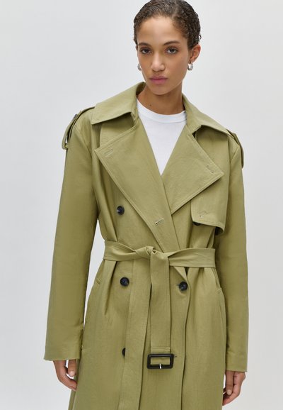 Γυναίκα με κοντά σγουρά μαλλιά φοράει ανοιχτό ελαιδί μπουφάν τύπου trench με ζώνη πάνω από λευκή μπλούζα, στέκεται μπροστά σε απλό φόντο.