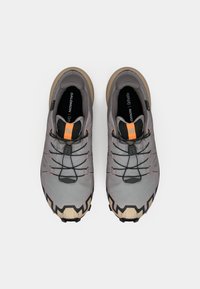 Graue Sportschuhe mit einer strukturierten Oberseite, elastischen Schnürsenkeln und schwarzen Akzenten. Sie verfügen über eine dicke, robuste Sohle und eine auffällige orangefarbene Markierung.