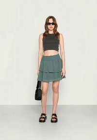 Top curto cinza, saia verde em camadas, mala preta de ombro e sandálias pretas chunky. Óculos de sol elegantes completam o visual.