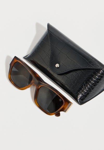 Καφέ γυαλιά ηλίου από καμηλοπάρδαλη με σκούρους φακούς δίπλα σε μαύρη θήκη από συνθετικό δέρμα κροκόδειλου με κούμπωμα πατιλού.