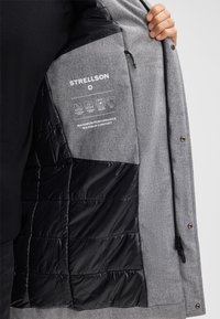 Graue Jacke mit einem schwarzen isolierten Futter, das ein Logo und Pflegehinweise innen aufgedruckt hat, sowie einem Reißverschluss mit Druckknöpfen.