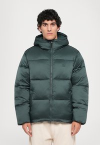 Mann in einer dunkelgrünen gepolsterten Winterjacke mit Kapuze und Frontreißverschluss, kombiniert mit hellbeigen Hosen, blickt nach vorne.