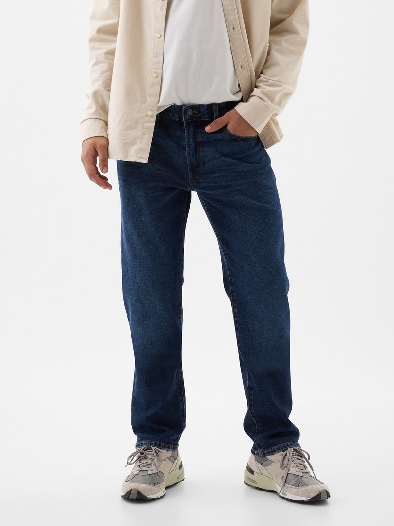 Homme portant un jean bleu foncé, une chemise beige à boutons par-dessus un t-shirt blanc, et des baskets grises, se tenant avec une main dans la poche.