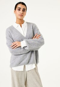 Grauer Strickpullover mit einem V-Ausschnitt, strukturiertem Stoff und lockerer Passform, über einem weißen Hemd mit Knöpfen getragen und kombiniert mit hellen, gefalteten Hosen.