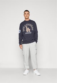 Sweatshirt marine avec "Los Angeles Dodgers" imprimé en beige, associé à un pantalon de survêtement cargo gris. Des baskets blanches complètent la tenue.
