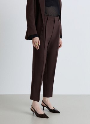 Persona che indossa pantaloni sartoriali marrone scuro, blazer abbinato, top nero in rete, anelli d'oro e scarpe slingback nere con tacco basso.