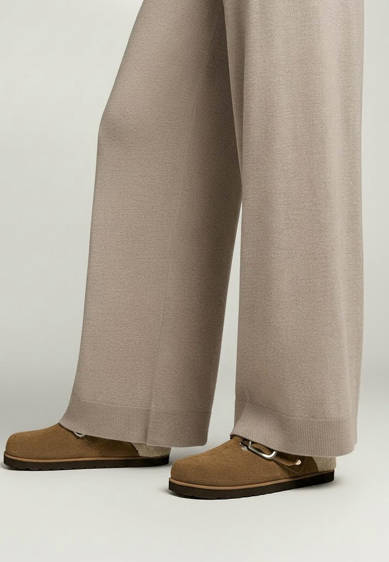 Béžové nohavice s širokými nohavicami z mäkkého pleteného materiálu, skombinované s hnedými semišovými slip-on topánkami s kovovou prackou.
