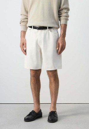 Homme portant un pull beige à manches longues, un short blanc jusqu'aux genoux avec une ceinture noire, et des mocassins en cuir noir, debout sur un sol gris.
