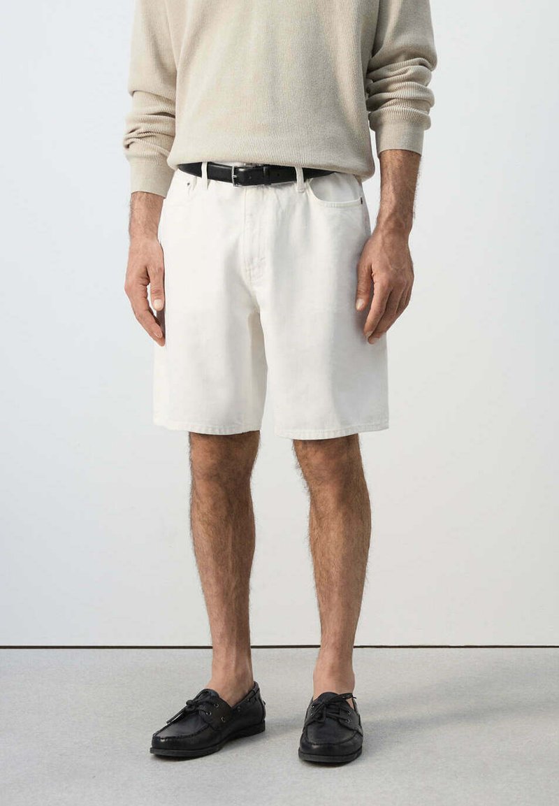 Homme portant un pull beige à manches longues, un short blanc jusqu'aux genoux avec une ceinture noire, et des mocassins en cuir noir, debout sur un sol gris.