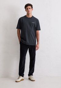 Ciemnoszary bawełniany t-shirt z krótkim rękawem i luźnym krojem, zestawiony z ciemnymi sztruksowymi spodniami i białymi trampkami z beżowym akcentem.