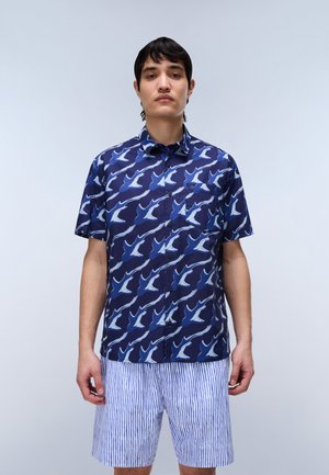 Ung mand står iført en marineblå kortærmet skjorte med hvide fugle og lyseblå og hvide kortstribede shorts mod en ensfarvet baggrund.