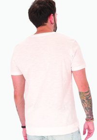 Uomo con capelli scuri e tatuaggio floreale sulla spalla, che indossa una maglietta bianca a maniche corte e jeans azzurri chiari, di spalle.