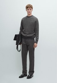 Maglione grigio a maglia con scollo a girocollo, abbinato a pantaloni grigio scuro. La modella tiene in mano una borsa in pelle nera, in piedi su uno sfondo chiaro.