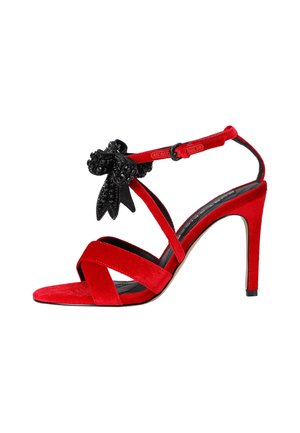 Sandalo con tacco alto in suede rosso, caratterizzato da più cinturini incrociati, decorato con un accento di fiore nero e dotato di una fascia alla caviglia con fibbia.