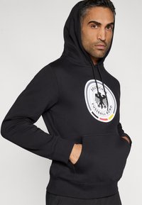 Fekete kapucnis pulóver elülső zsebbel, kerek logóval, amelyen egy sas és a "Deutscher Fussball-Bund" felirat látható. A kapucni húzózsinórokkal ellátva.