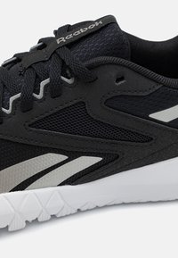 Gros plan sur une chaussure de sport Reebok noire avec semelle blanche, panneaux en maille et accents argentés réfléchissants sur le côté et la languette.