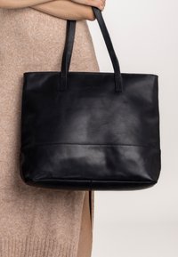 Crna kožna torba sa dvije ručke, s glatkom teksturom, minimalističkim dizajnom i prošivenim detaljem na dnu.