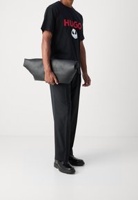 Mustat Hugo T-paita punaisella logolla, yksiväriset mustat housut ja tyylikäs musta nahkakotelo, jota pidetään yhdessä kädessä. Minimalistinen muotoilu ja rakenne.