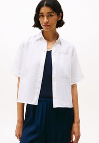 White linen short-sleeve shirt with a button-down front, chest pocket, and slightly cropped design, layered over a dark tank top.