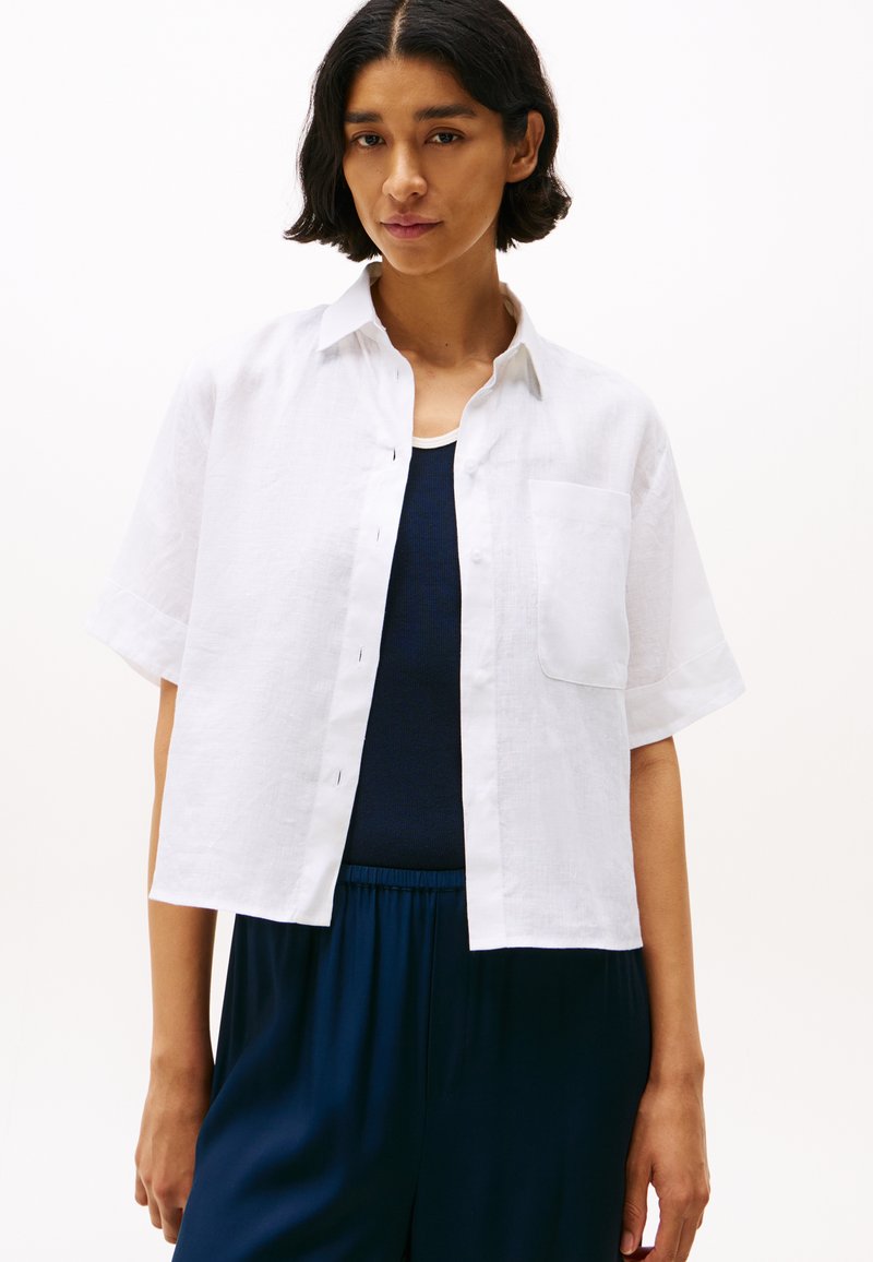 White linen short-sleeve shirt with a button-down front, chest pocket, and slightly cropped design, layered over a dark tank top.