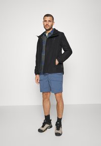 Veste noire imperméable avec capuche, associée à une chemise à carreaux grise et un short cargo bleu. Portée avec des chaussures de randonnée grises et noires.