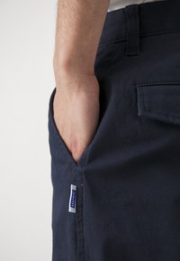 Mørkeblå bomullsbukser med en sidelomme, som har et lite hvitt merke med blå tekst. Stoffet har en teksturert overflate.