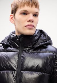 Zwarte pufferjack met een glanzende afwerking, voorzien van een hoge kraag, een voorrits en een subtiele Calvin Klein-logo op de borst.