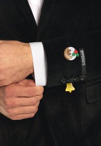 Man justerar svarts kostymsmanschetten med skjortärmen i vitt, med broderade julpudding- och klockpatcher och "OPPOSUITS"-etikett.