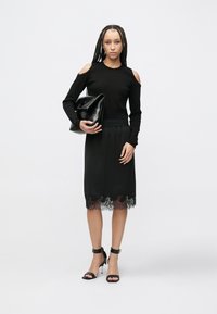 Pulover negru cu mâneci lungi și umeri decupați, asortat cu o fustă neagră midi cu dantelă la margine. Modelul poartă o geantă neagră și pantofi cu toc negri.