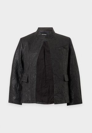 Femme aux cheveux courts portant une veste noire texturée, un t-shirt blanc imprimé, un pantalon large en cuir noir, des chaussures noires, tenant un sac à main noir.