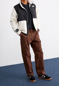 Fleecejacke in Schwarz und Creme mit Reißverschluss vorne, kombiniert mit braunen Cargo-Hosen mit Taschen und lockerer Passform. Schwarze Sneakers mit Gummisohlen.