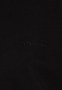 Schwarzer Stoff mit einem gestickten Logo "Smilodex" in schwarfem Faden, das eine glatte Textur und ein minimalistisches Design zeigt.