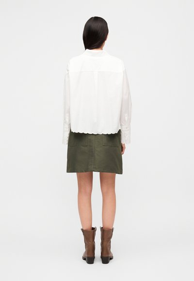 Λευκό μπλουζάκι με μακριά μανίκια, με κυματιστό τελείωμα και λεπτομερείς μανσέτες, συνδυασμένο με πράσινη φούστα και καφέ μποτάκια με τακούνι.