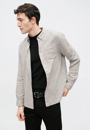 Chemise gris clair à boutons avec une finition texturée, poche sur la poitrine et coupe décontractée, portée sur un pull noir et un pantalon sombre. Ceinture noire incluse.