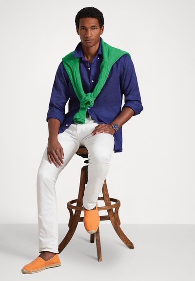 Μοντέλο με σκούρο μπλε πουκάμισο με κουμπιά, λευκό παντελόνι και πορτοκαλί εσπαντρίγιες, με ένα πράσινο πουλόβερ περασμένο γύρω από τους ώμους.