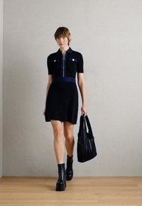 Robe noire à manches courtes avec fermeture éclair à l'avant, col contrastant et ceinture. Le mannequin tient un sac à main noir texturé et porte des bottes noires épaisses.